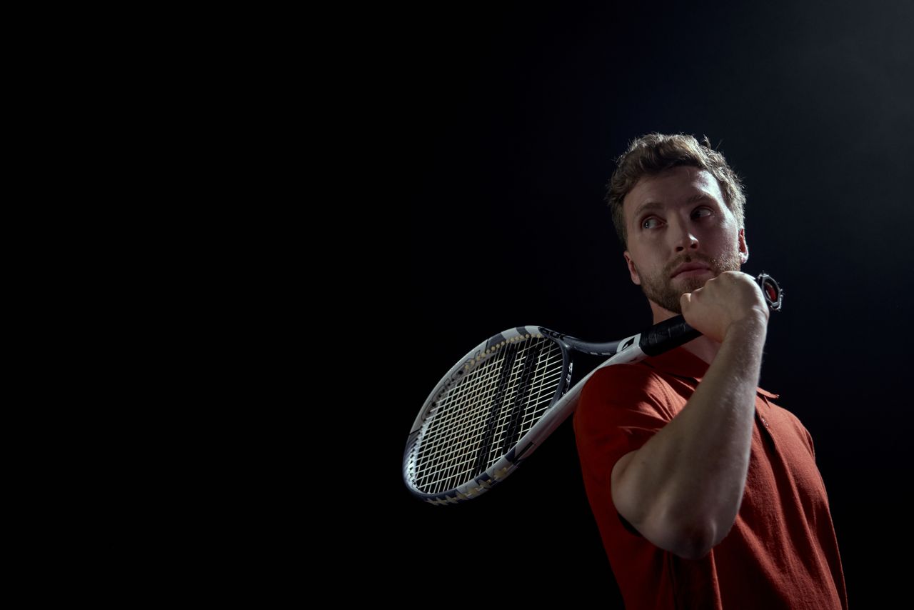 Services A thoughtful male tennis player holding a racket, set against a dark background.